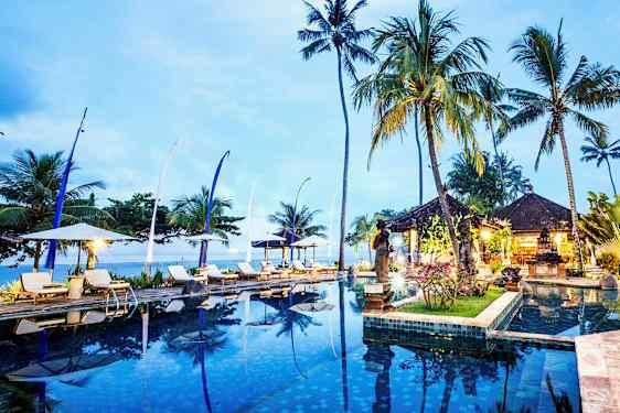 Et infinitybasseng omgitt av solsenger, parasoller, høye palmer og et åpent paviljongområde ved skumring. (AI-generert tekst)