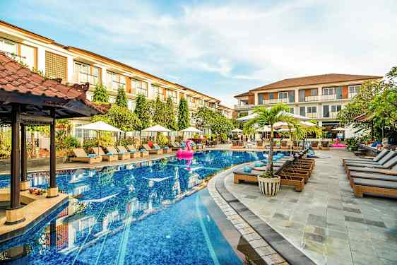 Et hotellbasseng med solsenger og parasoller rundt omkring, samt noen bygninger i bakgrunnen. (AI-generert tekst)