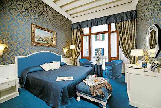 Et vakkert innredet hotellrom med blå og gyldne detaljer, stor seng, sittegruppe og store vinduer. (AI-generert tekst)