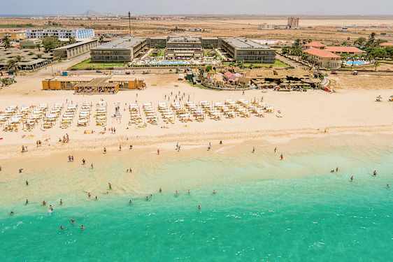 Luftfoto av en strand med turkis vann, solsenger og parasoller, samt bak en hotellbygning. (AI-generert tekst)