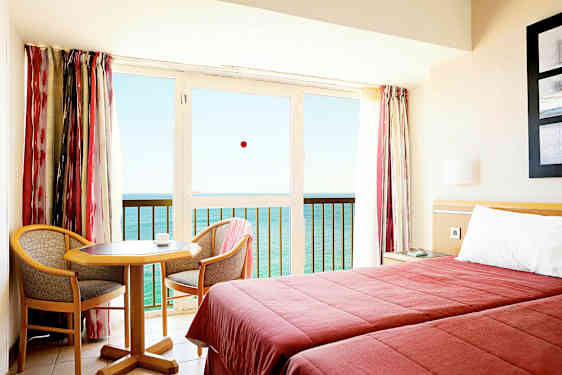 Et hotellrom med to senger, et lite bord og to stoler, samt en balkong med utsikt over havet. (AI-generert tekst)