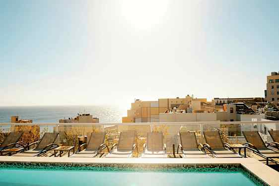 Et basseng med solsenger foran en utsikt over havet og bygninger. (AI-generert tekst)