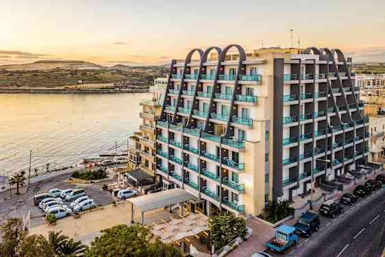 En bygning ved vannet med moderne etasjer og balkonger. I forgrunnen sees parkerte biler og en vei. (AI-generert tekst)