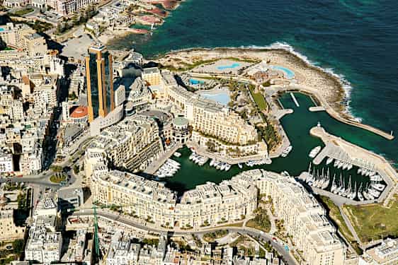 Flyfoto av en kystby med høye bygninger, småbåthavn og havet i bakgrunnen. (AI-generert tekst)