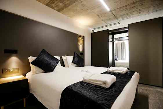 Soverom med en moderne dobbeltseng, hvitt sengetøy og mørke dekorative puter. Et nattbord med myk belysning står ved siden av. (AI-generert tekst)