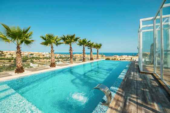 Et utendørs basseng med turkist vann, omgitt av palmer og med utsikt over en by og havet. (AI-generert tekst)