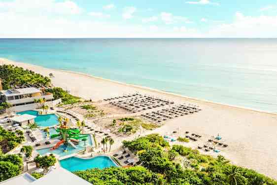 Bilde av en sandstrand ved havet med blått vann. På stranden er det solsenger og parasoller. I forgrunnen sees et basseng med vannsklier samt omkringliggende grønne planter. (AI-generert tekst)