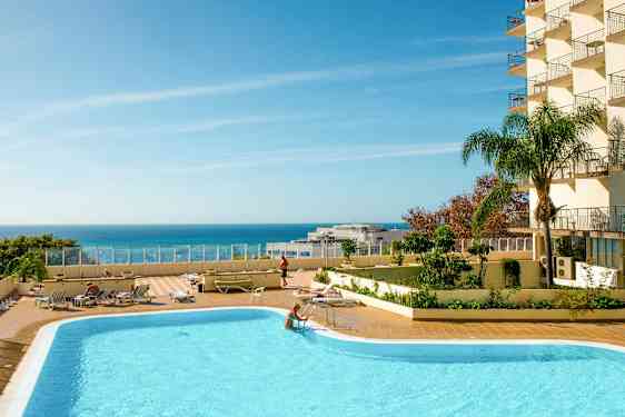 Et utendørs basseng med solsenger og en klar blå himmel over havet. (AI-generert tekst)