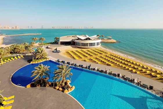 Et bilde av et moderne hotellkompleks ved havet. I forgrunnen sees et stort basseng og solsenger på en strand. (AI-generert tekst)