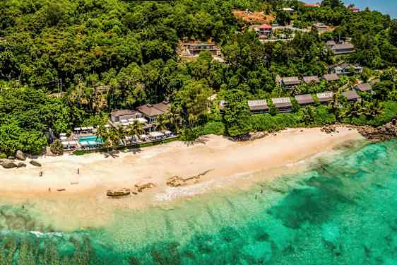 Strand med turkis vann, omgitt av frodig vegetasjon og bygninger. (AI-generert tekst)