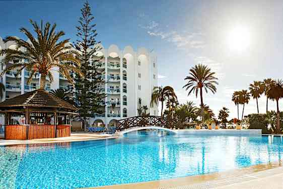 Et utendørs basseng ved et hotell, med palmer og en trebro over bassenget. (AI-generert tekst)