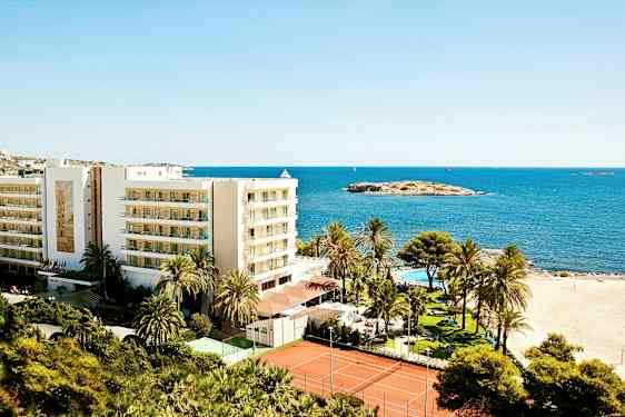 Bilde av et feriested ved stranden med en bygning, palmer, basseng og tennisbaner, lokalisert ved sjøen. (AI-generert tekst)