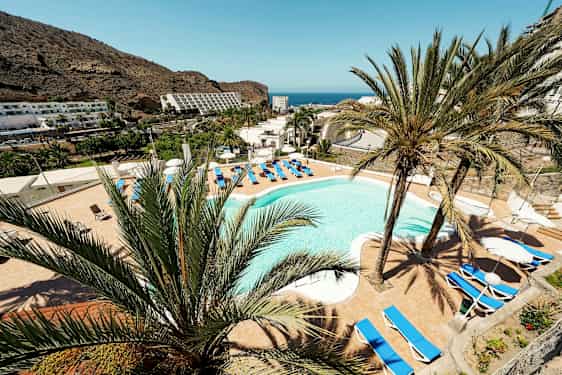 Utsikt over et hotellbasseng omgitt av solsenger og palmer med fjell og hav i bakgrunnen. (AI-generert tekst)