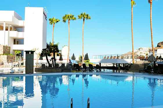 Et basseng omgitt av solsenger, parasoller og palmer, med en bygning og klar blå himmel i bakgrunnen. (AI-generert tekst)