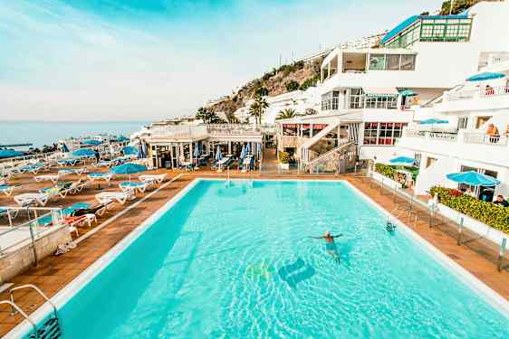 Bilde av et stort utendørs basseng omgitt av solsenger og parasoller med hvite bygninger og hav i bakgrunnen. (AI-generert tekst)