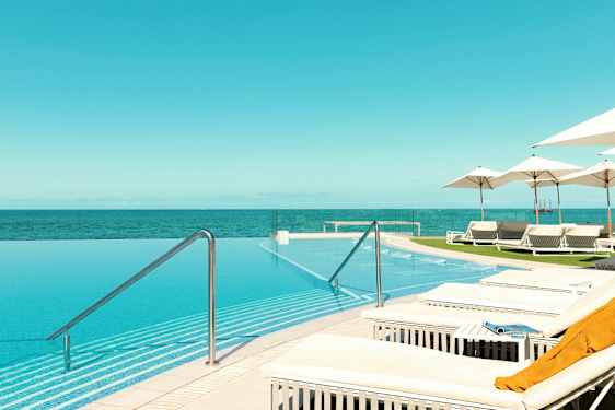 Et infinity-basseng med utsikt over havet, omgitt av solsenger og parasoller. (AI-generert tekst)