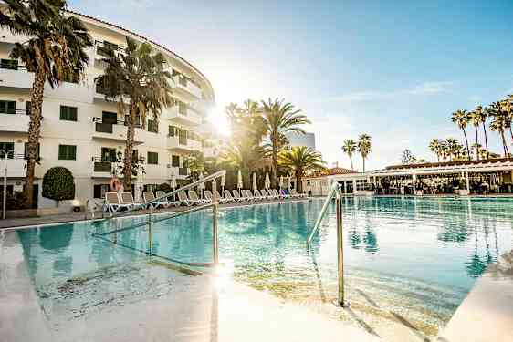 Et utendørs basseng foran et hotell omgitt av palmer og solsenger. (AI-generert tekst)