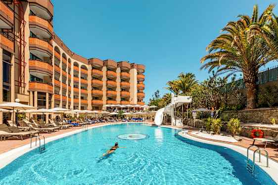 Et svømmebasseng med en kvinne som svømmer, omgitt av solsenger og et hotell i bakgrunnen. (AI-generert tekst)