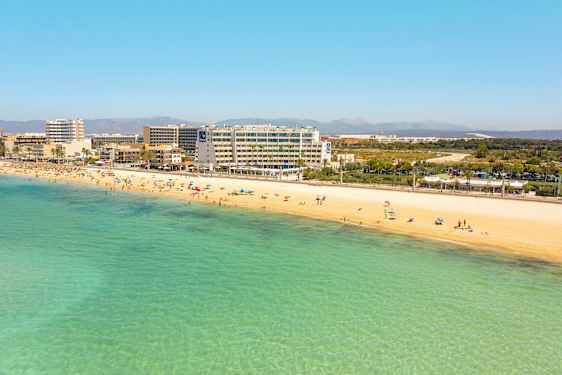 Sandstrand med badende personer og klart turkisblått vann. Hoteller og bygninger i bakgrunnen. (AI-generert tekst)