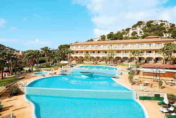 Bildet viser et hotell med et stort basseng foran og en omkringliggende terrasse med solsenger. Bakgrunnen har en frodig bakke og klar blå himmel. (AI-generert tekst)