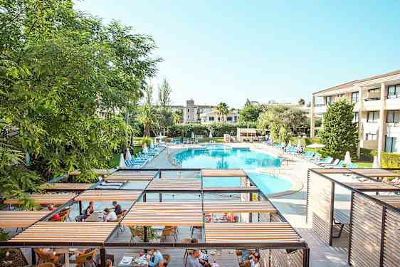 Et feriested med et stort ovalt basseng omgitt av solsenger og parasoller, og et spiseområde med pergolaer. (AI-generert tekst)