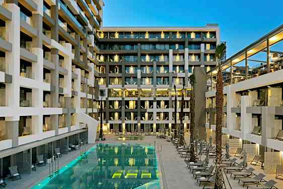 En moderne hotellbygning med balkonger og et opplyst basseng omgitt av solsenger. (AI-generert tekst)