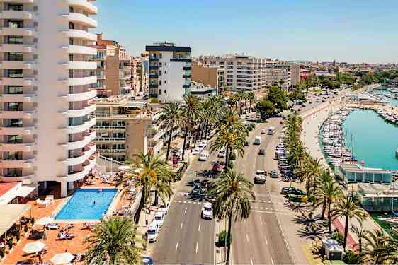 Bilde som viser en gate med palmetrær, et basseng ved et bygningskompleks og en havn med båter. (AI-generert tekst)