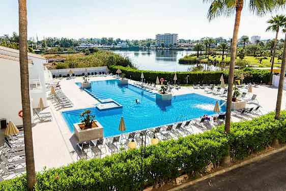 Utendørs svømmebasseng med solsenger og parasoller rundt, omgitt av grønne planter og med utsikt over en innsjø og et åpent landskap. (AI-generert tekst)