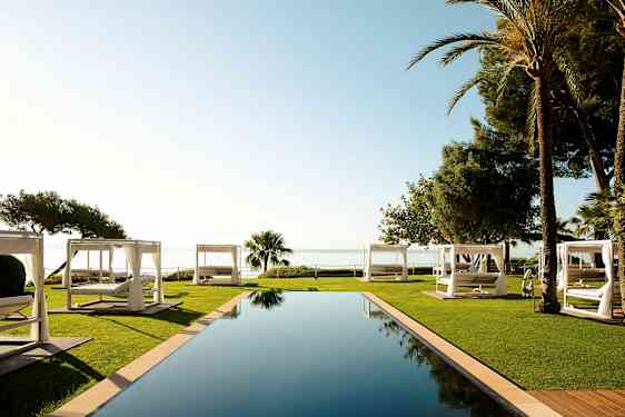 Et infinitybasseng omgitt av plen og hvite solsenger med sjøutsikt. (AI-generert tekst)