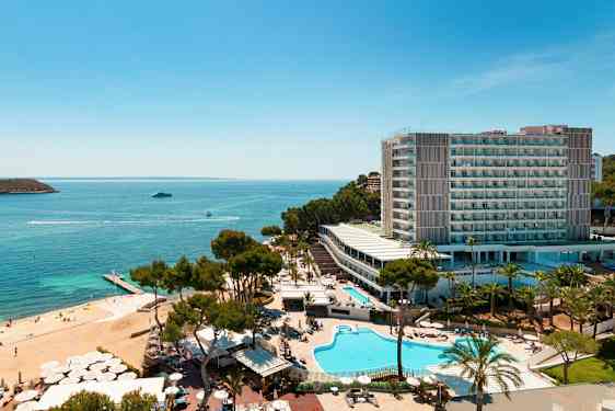 Flyfoto av et hotellkompleks ved sjøen med bassenger, grøntområder og en strand. (AI-generert tekst)