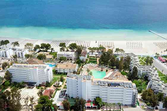 Luftfoto av et hotellområde nær stranden med parasoller på sanden og en klarblå sjøutsikt. (AI-generert tekst)