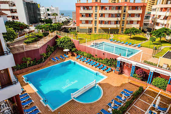 Et hotellområde med to bassenger, solsenger og omkringliggende bygninger. (AI-generert tekst)