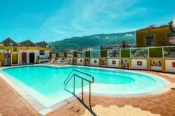 Utendørs basseng med solsenger i en solrik setting, omgitt av bygninger og med fjell i bakgrunnen. (AI-generert tekst)