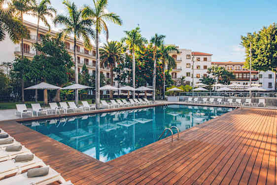 Et utendørsbasseng omgitt av solsenger, parasoller og frodig vegetasjon, med en bygning i bakgrunnen. (AI-generert tekst)