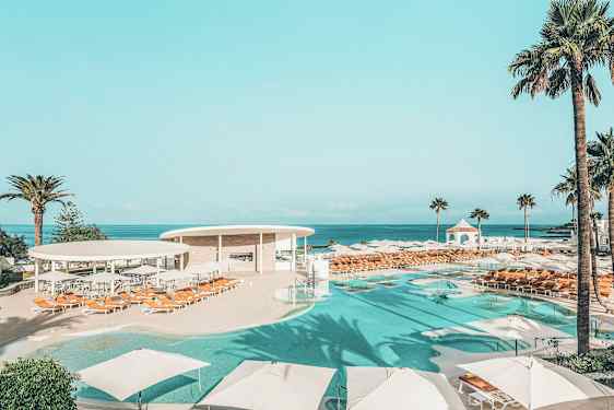 Et bassengområde med parasoller og solsenger, omgitt av palmer og med utsikt over havet. (AI-generert tekst)