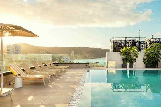 Utsikt over et basseng med solsenger og parasoller, i bakgrunnen sees en by og havet. (AI-generert tekst)