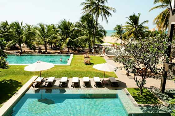 Bildet viser et hotellbassengområde med solsenger og parasoller, omgitt av tropiske palmer, med utsikt mot stranden og havet. (AI-generert tekst)