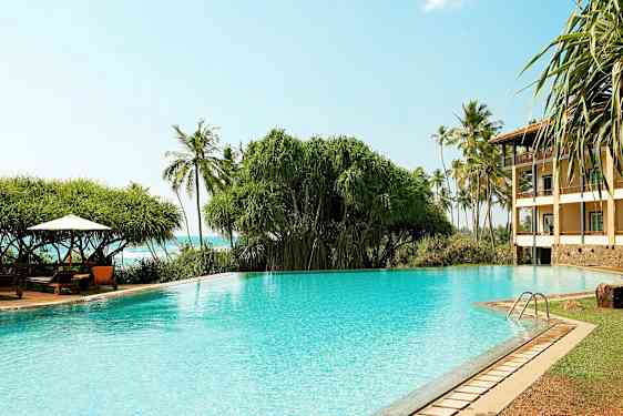 Et krystallklart svømmebasseng med vegetasjon og palmer i bakgrunnen samt en bygning til høyre. (AI-generert tekst)