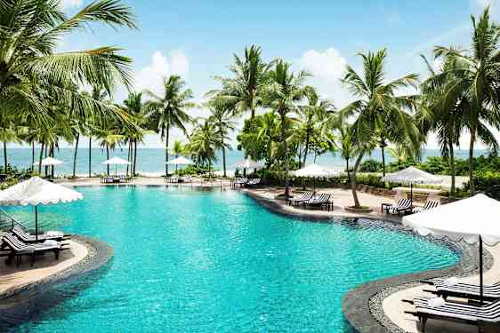 Bilde av et svømmebasseng omgitt av palmer og solsenger med parasoller ved havet. (AI-generert tekst)