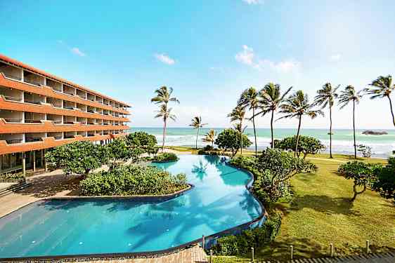 En hotellbygning ved stranden med et stort basseng, palmer og havet i bakgrunnen. (AI-generert tekst)