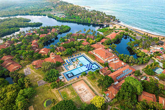 Luftfoto av et feriested med terrakottafargede tak, et stort basseng og omkringliggende grøntområder. (AI-generert tekst)