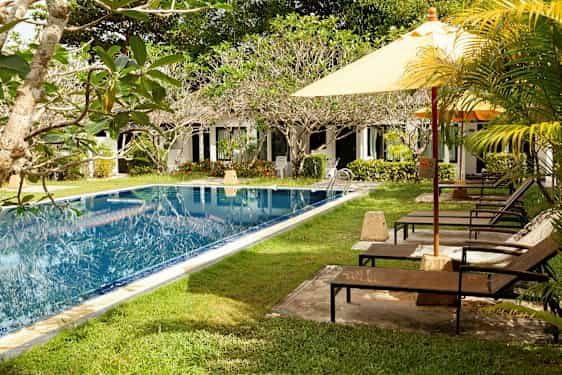 Et basseng med grøntområder og solsenger ved siden av, samt en rekke hvite bygninger i bakgrunnen. (AI-generert tekst)