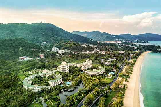 Luftfoto av et kystområde med hotellbygninger, omkringliggende grøntområder og en strand. (AI-generert tekst)