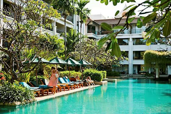 Bilde av et utendørs basseng omgitt av solsenger, parasoller og planter, med en bygning i bakgrunnen med balkonger. (AI-generert tekst)