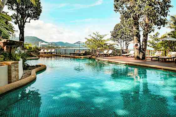 Utendørsbasseng med klart vann omgitt av solsenger, parasoller og grønn vegetasjon. Utsikten viser fjell og hav i bakgrunnen. (AI-generert tekst)