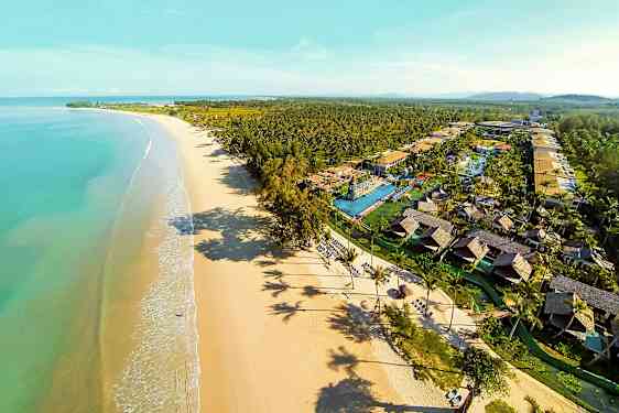 Flyfoto av en lang sandstrand med turkis vann og en ferieanlegg med bungalower og bassenger. (AI-generert tekst)