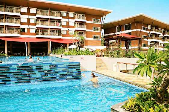 Et hotell med to etasjer er synlig bak et utendørsbasseng hvor mennesker svømmer. Grønne planter og terrasseområder omgir bassengområdet. (AI-generert tekst)