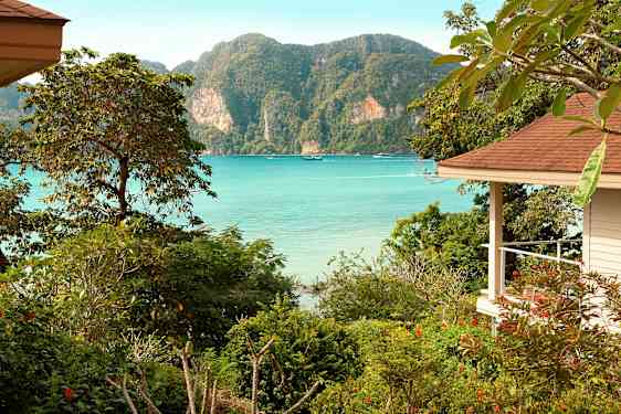 En utsikt over en havsbukt med klart blått vann, omgitt av frodig vegetasjon og fjell, med en del av en bygning til høyre. (AI-generert tekst)