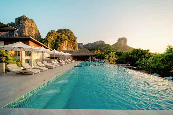 Et basseng med solsenger og parasoller ved en resort omgitt av en naturskjønn atmosfære med fjell i bakgrunnen. (AI-generert tekst)