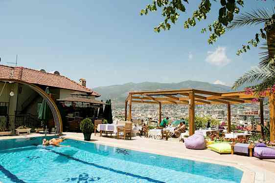 Et utendørs basseng ved et hotell med solsenger og parasoller. En trepergola og utsikt over en by og fjell i bakgrunnen. (AI-generert tekst)